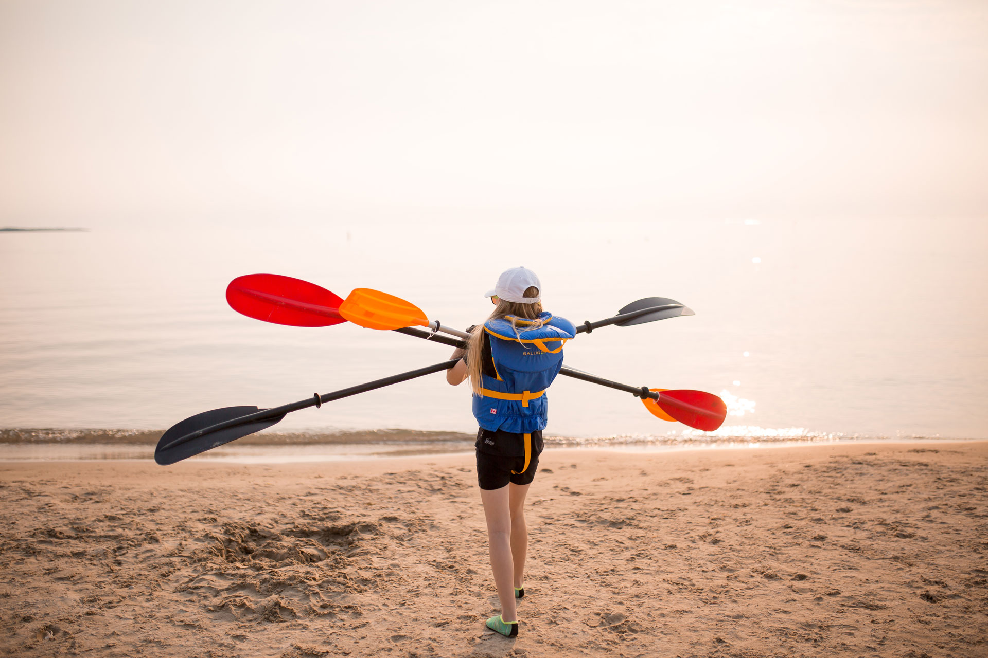 girl wearing a life jacket and white hat on a beach walking towards water holding oars